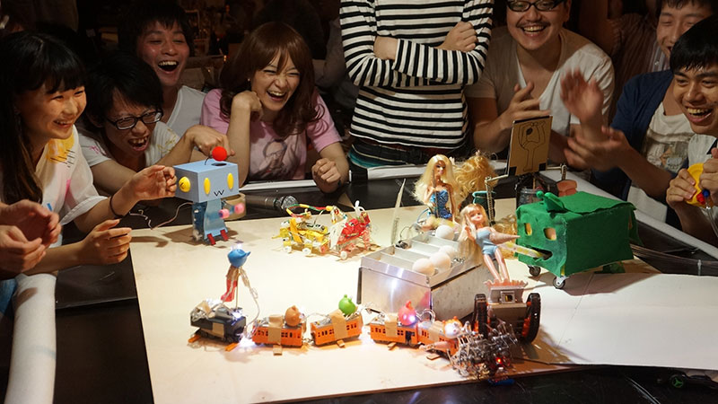 A crowd laughing and cheering around a tabletop robot battle
