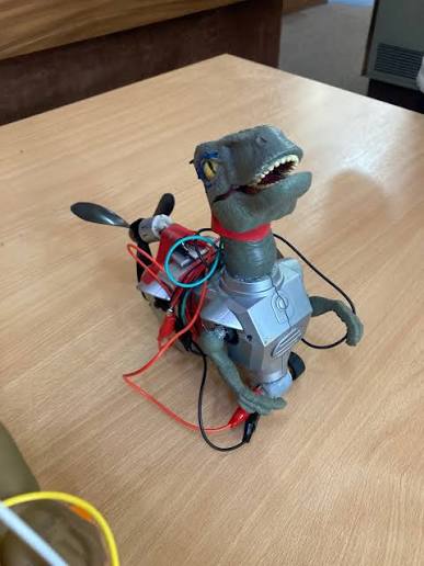 A toy dinosaur strapped to a motorised toy car base with loose wires